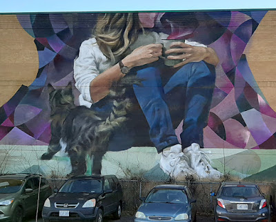 Large outdoor wall mural of a young woman sitting on a ledge, holding a coffee mug in two hands. A small cat stands looking at her. Her face is not included in the mural - it's as if they ran out of building height.