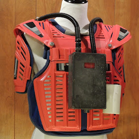 Front view of the restored laser tag pack displayed on a dressmaker's dummy. It is red in color with a dull-colored aluminum box on the front, and a white holster tube on the side. Cables go up and over the shoulders to the back. There is no gun.