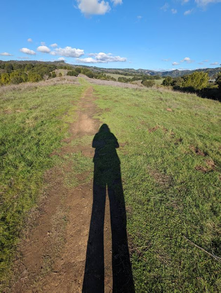 Photo of a person's long shadow on a path at the top of a grassy hill, with blue sky and more hils in the background.