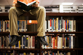 young man sitting on top of bookshelves reading a book