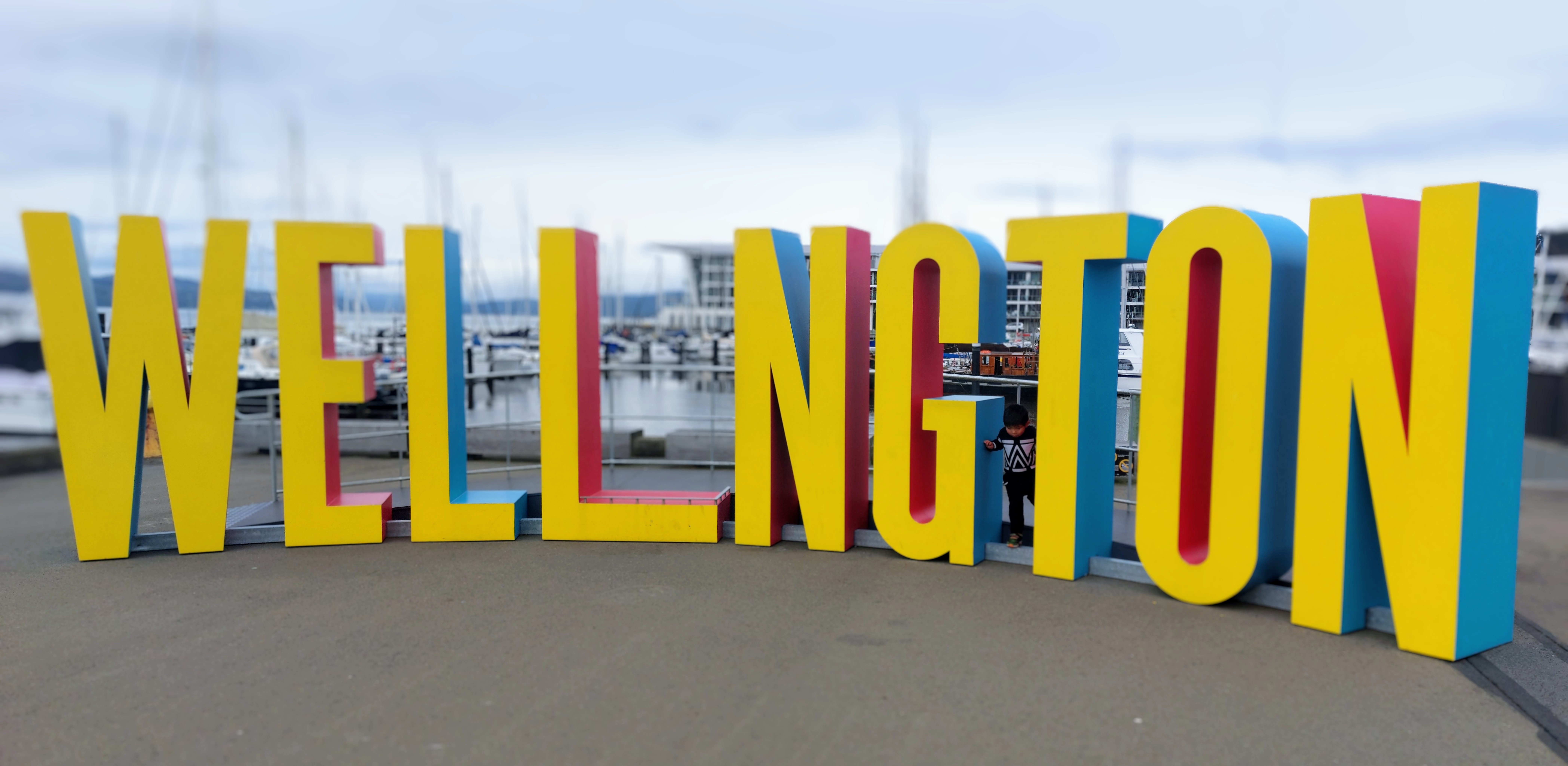 Yellow (with red and blue edges) WELL_NGTON interactive tourist sign front and centre. Large people sized letters with space for someone to be the 'I'