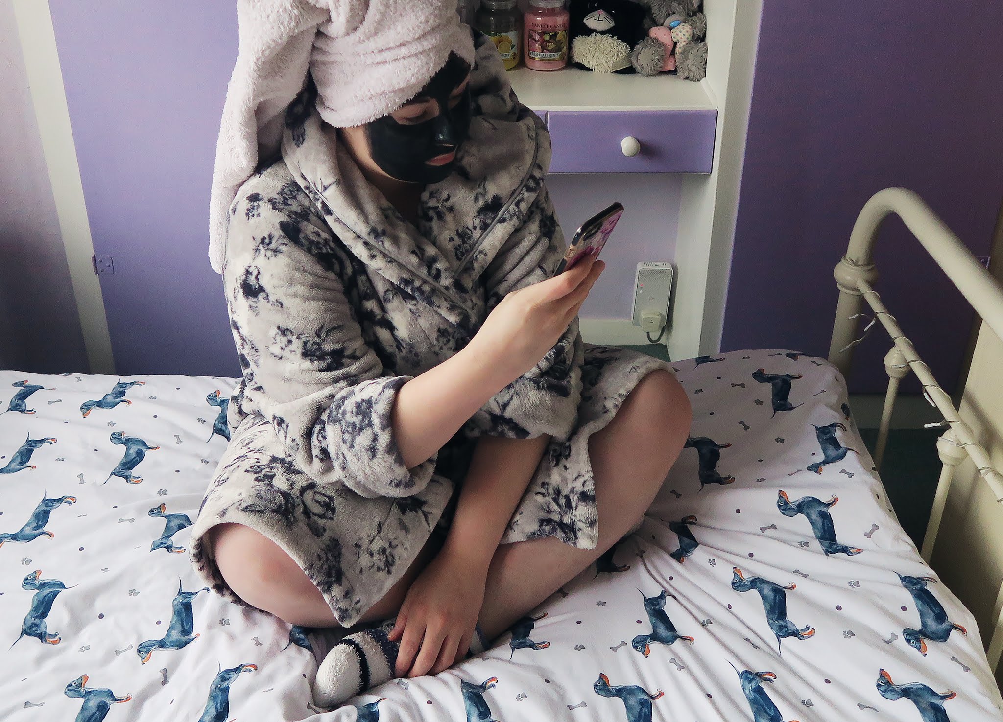 Grace is sitting on the bed in her dressing gown. Her hair is wrapped in a towel and she has a charcoal facemask on