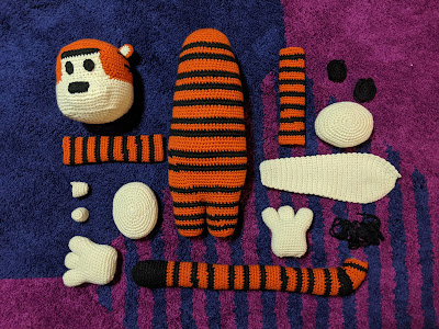 A flat lay of the parts of crochet Hobbes (tiger) plushie, where all the body parts are laid out before construction. They are on a purple and blue geometric carpet.