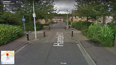 A view of a street where the road is narrowed at the place a cycle track crosses from each side. there is a footway on both sides with bushes behind those. The paving changes type and colour at the junction.