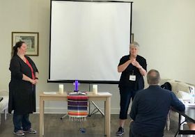 During the SEJ of The UMCD business meeting, Betty Ostrom, the new spiritual advisor of the SEJ board, standing next to the altar, prays for the new board, while Mary Ann Deters (board president) looks on, and Rev. Yates (voice) interprets the prayer.