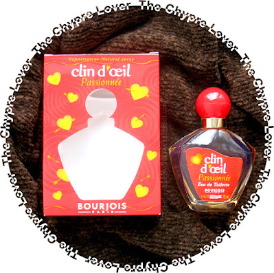 The pentagonal bottle and red cardboard box of Clin d'Oeil Passionnee by Bourjois (eau de toilette) lying flat on a brown, thick, textured sweater