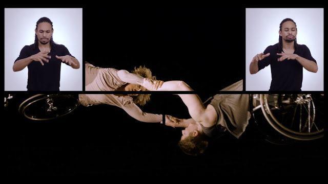 Image Description: A black-lined grid overlays an image of Alice and Laurel outstretched in full horizontal flight. Alice, a multiracial Black woman with short curly hair and coffee-colored skin, and Laurel, a pale white woman with short-cropped brown hair, twist and connect in a black sky. Their hands link to each other’s wrists to form a human infinity loop in midair: Laurel on her back and Alice twisting sideways. Their wheelchairs glint in the light. Brandon, a mixed-race Black artist with long braids half tied up, signs in the top corners of the screen. Alice Sheppard and Laurel Lawson of Kinetic Light; still from One + One Make Three/Safety Third Productions.