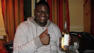 Man in a grey and white striped hooded sweatshirt holding up his right hand in the thumbs up pose and a bottle of white wine standing upright in the palm of his left hand