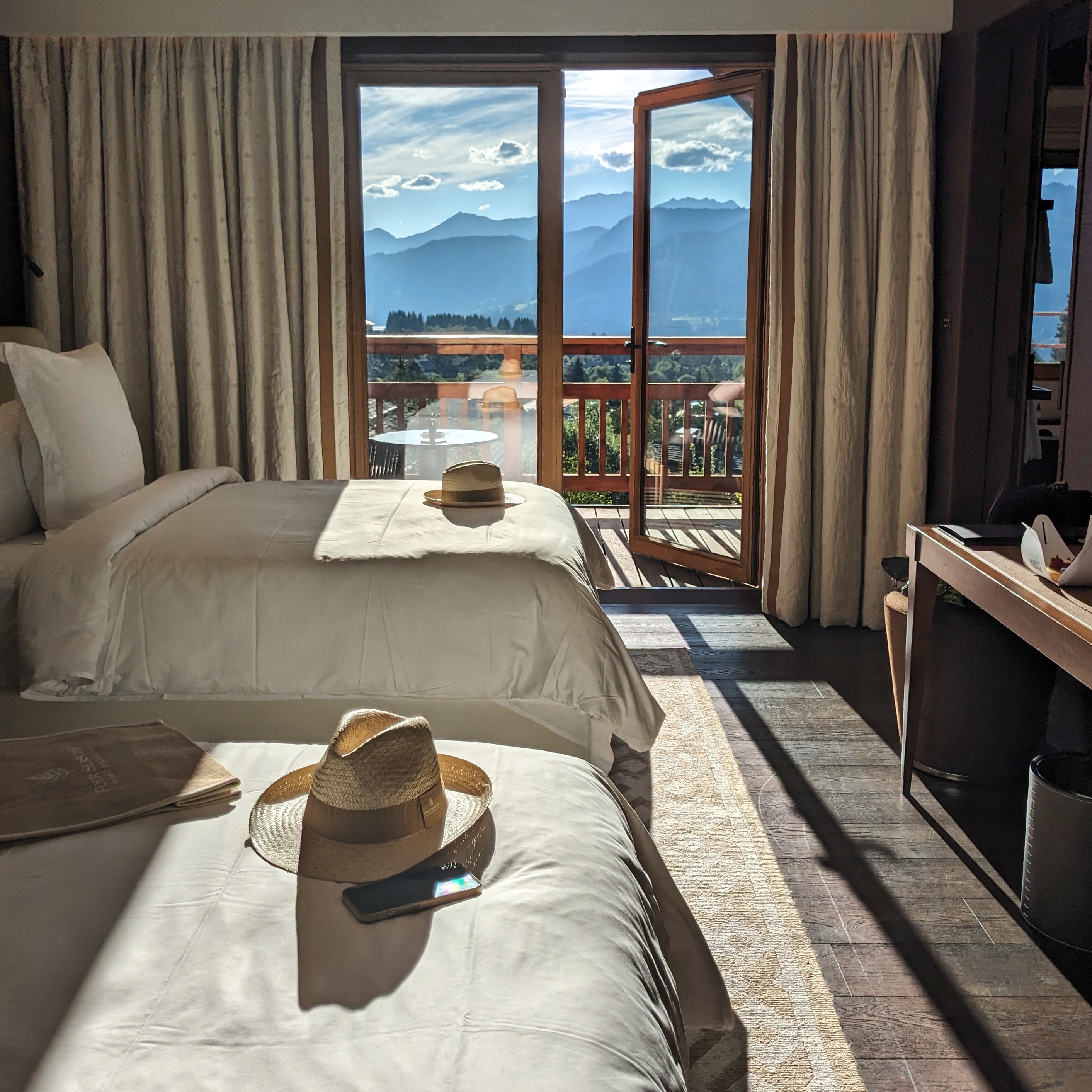 twin beds with panama hats on them in a deluxe twin bedroom at four seasons megeve with stunning mountain views seen through open doors