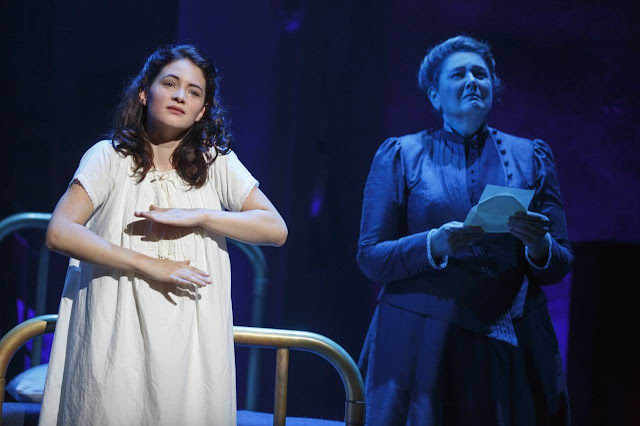 Sandra Mae Frank signando en la obra de teatro Spring Awakening con un camisón. A su lado, una mujer mayor con expresión triste y una carta en la mano