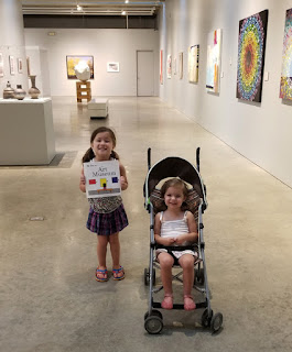 Two young girls smiling in an art gallery, one in a stroller, the other holds a children's book about visiting an art museum.