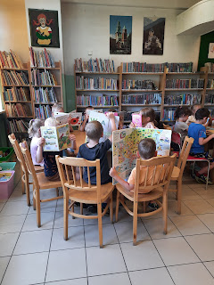 Fotografia przedstawia dzieci siedzące przy stoliku i oglądające książki. W tle znajduje się regał biblioteczny z książkami.