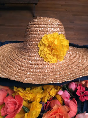 A straw hat with medium-blue fabric peeping out from under the brim, and a single bright-yellow chrysanthemum attached at the front intersection between the crown and brim. The hat is sitting on a pile of bright-yellow, peach, coral, and pale-pink fake flowers. 