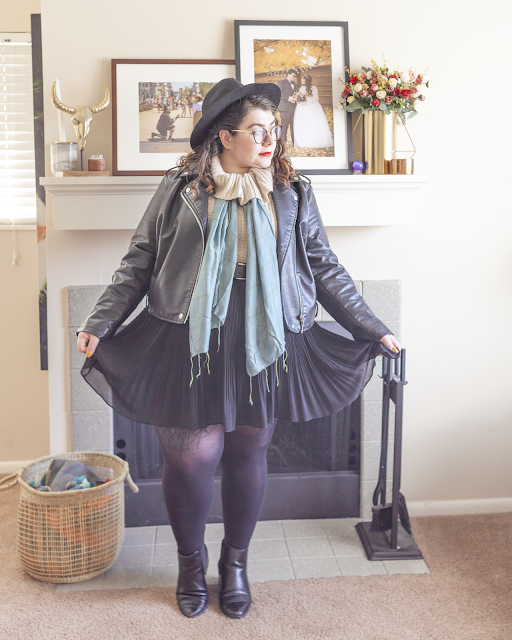 An outfit consisting of a wide brim hat, black moto jacket over a cream cowl neck sweater with a seafoam colored scarf tied under the cowl, black pleated skirt and black boots.