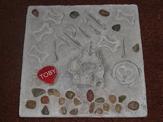 Actual picture of Toby's stone, this is when it's finished and out of the mold. It has his paw print in the middle, with his red name tag to the left, below the paw print his name is spelled out in rocks. In random places there are rocks & dog bone imprints.  It also says April 10 (2010)