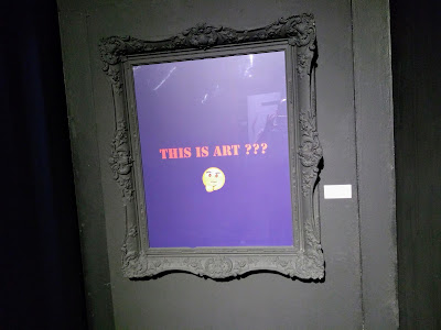 Quadro escrito "This is Art?" com um emoticon pensando