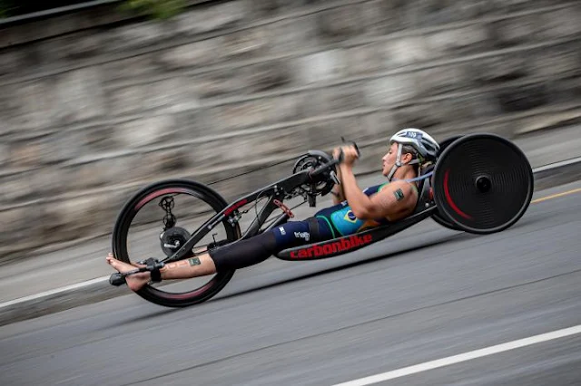 Jessica Messali deitada em sua cadeira adaptada e manuseando o guidão para pedalar. Ela é morena e branca