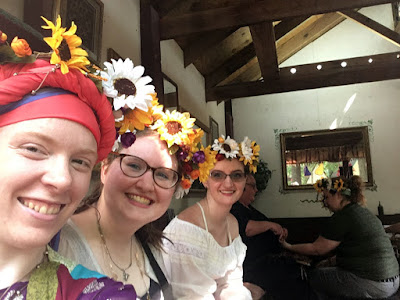 A selfie of three young white women in a row, all wearing colorful flower crowns and smiling at the camera. In the background, inside a plaster-and-beam building, another young woman in a green tshirt and flower crown is leaning forward with her arm outstretched for a henna artist to paint on.