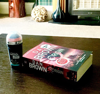 A small stubby black and silver rectangular bottle with a rounded top with a clear plastic lid with a silver label saying L'Oreal Mex Epxert Carbon Roll On in bright black font next to a rectangular book saying dan brown in white font on a brown rectangular table  on a bright background 