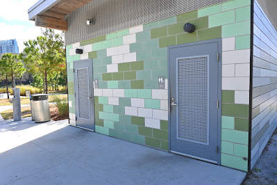 Two, unisex and accessible bathrooms are shown. They're in a small building, the outside of which is decorated with tile that is varying shades of green and white. To the left of the building are a few trashcans.