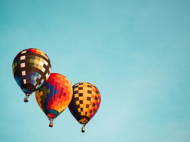 Three colorful hot air balloons float gracefully in the sky against a clear, light blue background, with ample negative space to the right suggesting movement and openness.