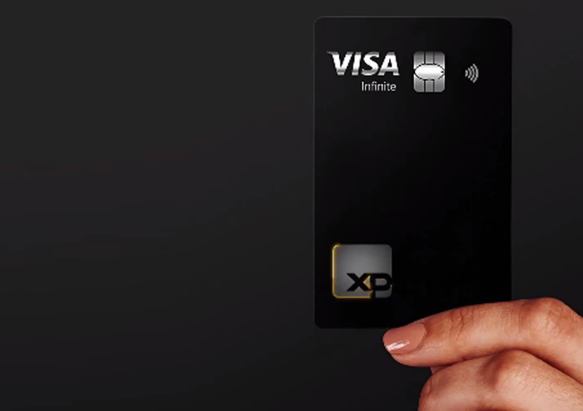 Imagem de fundo escuro mostra uma mão segurando um cartão de crédito XP Visa Infinite.