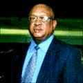 A color portrait of a heavy, middle-aged Black man in a suit and tie. He is bald and wearing eyeglasses.