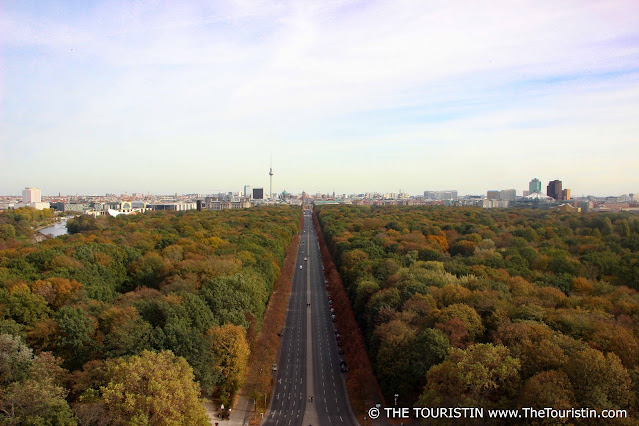 A long straight road leads through a large park, its trees in colourful autumn foliage and towards a city, under a soft blue sky.