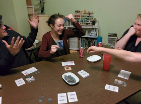 four players sitting around a table, looking animated at the current events of the game, playing Coup. There are cards and tokens on the table, with a bowl of spare tokens in the centre.