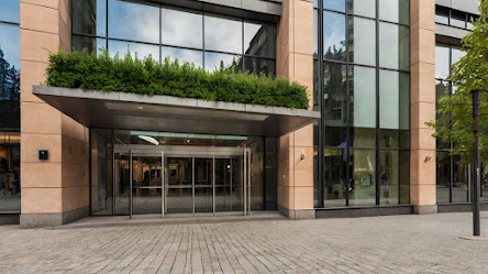 "Modern office building entrance with large glass doors, surrounded by reflective windows and a sleek architectural design. The entrance is adorned with greenery on top, set against a backdrop of trees and a clear sky."