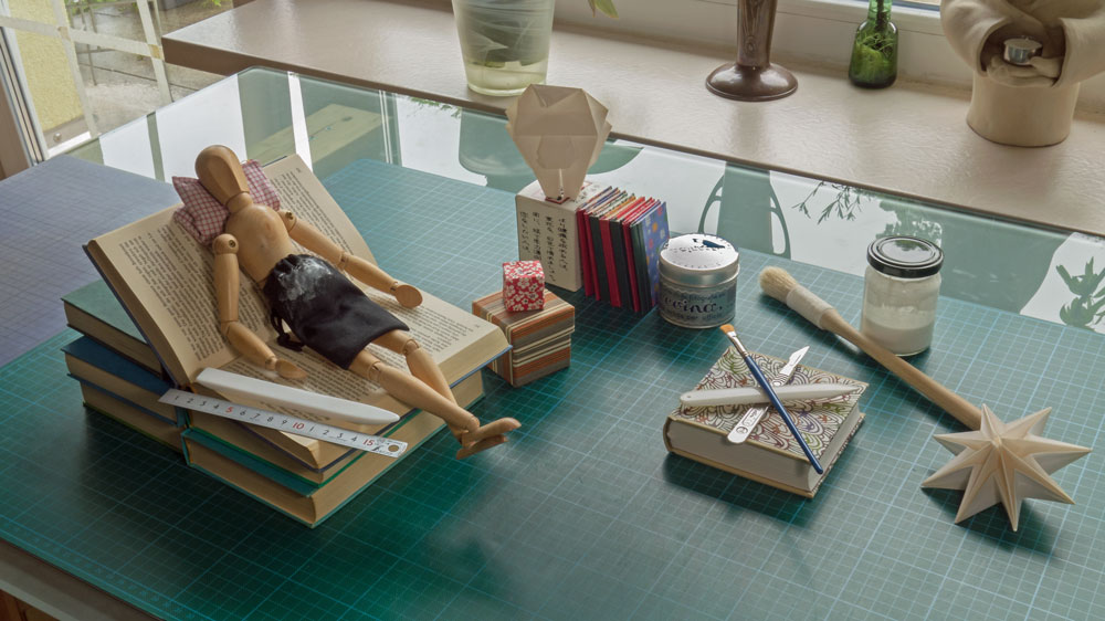 Gliederpuppe liegt auf einem aufgeschlagenen Buch wie auf einem Sofa, umgeben von Werkzeugen, kleinen Büchern und Origamimodellen