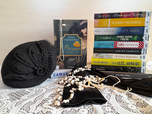 Książki ułożone w dwa stosy, obok czarny kapelusz, przed książkami czarne rękawiczki, biżuteria z pereł.