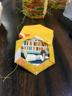 Close up of the back of a basted yellow hexagon, showing an illustration of a brunette girl lying on her stomach, reading a book in front of packed bookshelves. Stacks of finished yellow hexagons are just visible in the background.