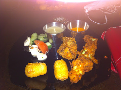 black plate with two mac and cheese bites, a chicken strip, fried zucchini, caprese salad and two dipping sauces.