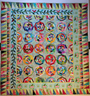 The spiderwebs in the center of the quilt are outlined in various single colors including red, green, blue, yellow, and purples. There is a narrow light green inner border followed by a border that is a printed toile on two sides and an applique vine on the other two. Finally there are two more borders of improvisational right triangles as the outer border.