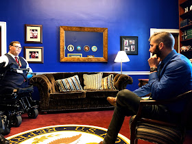 A man in a power wheelchair is speaking to a congressional staff member. They are in a congressional office. A rug underneath them bears a seal and says: "U.S. House of Representatives." The walls are dark blue.