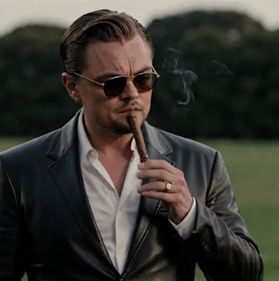 Leonardo DiCaprio with sunglasses on outside  from the waist up wearing a black leather blazer it's Titan slick and put in the cigar in his mouth