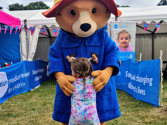 Image of a young girl in a tie dye top and plaited hair hugging a large character. The character is Paddington Bear, he is wearing his signature red hat and blue duffle coat. He is hugging the girl back. In the background is a tent and campaign banners advertising action medical research for children