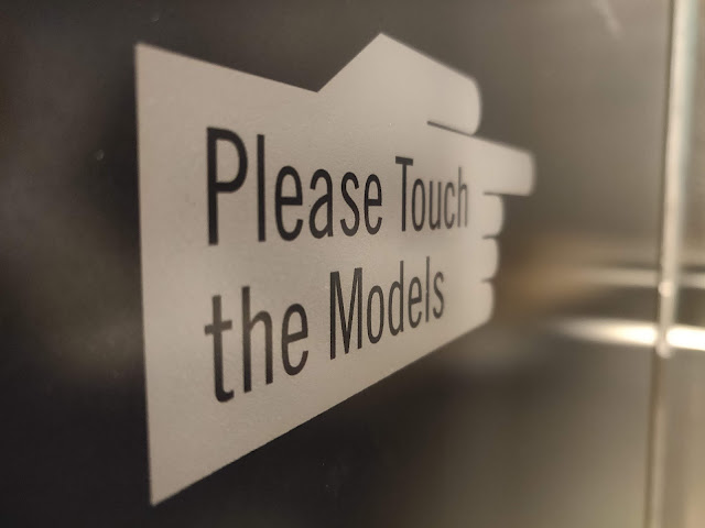 A sign in the shape of a hand with a pointing finger contains the words "please touch the models"