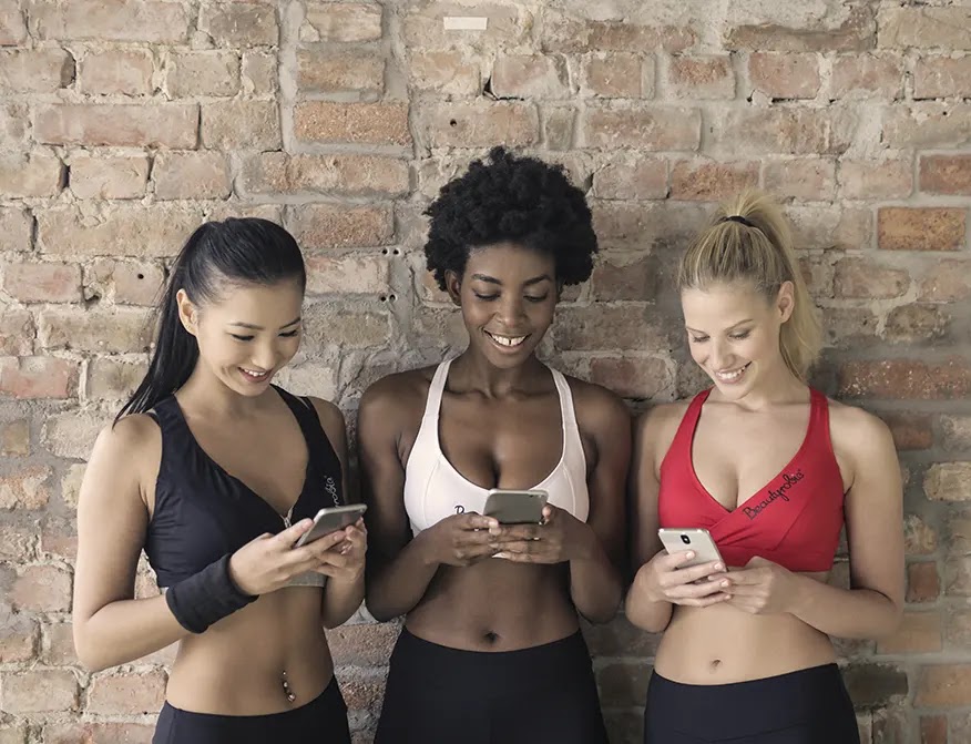 tres-mujeres-en-sujetadores-deportivos-de-varios-colores-sosteniendo-y-mirando-sus-telefonos-inteligentes