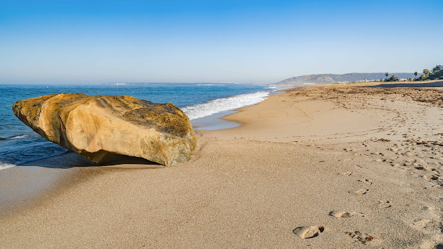 Playa de arena fina y llana con una gran roca en la orilla y las azules aguas del mar que la golpean.