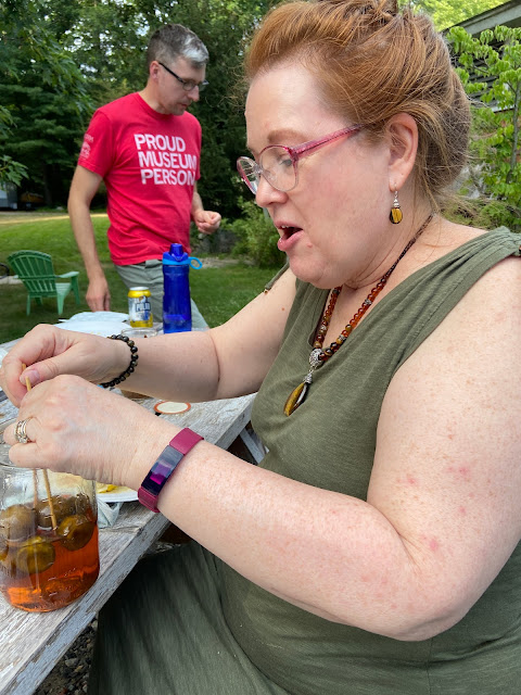 a red-haired woman wearing glasses uses two skewers to fish a pickled lime out of a mason jar