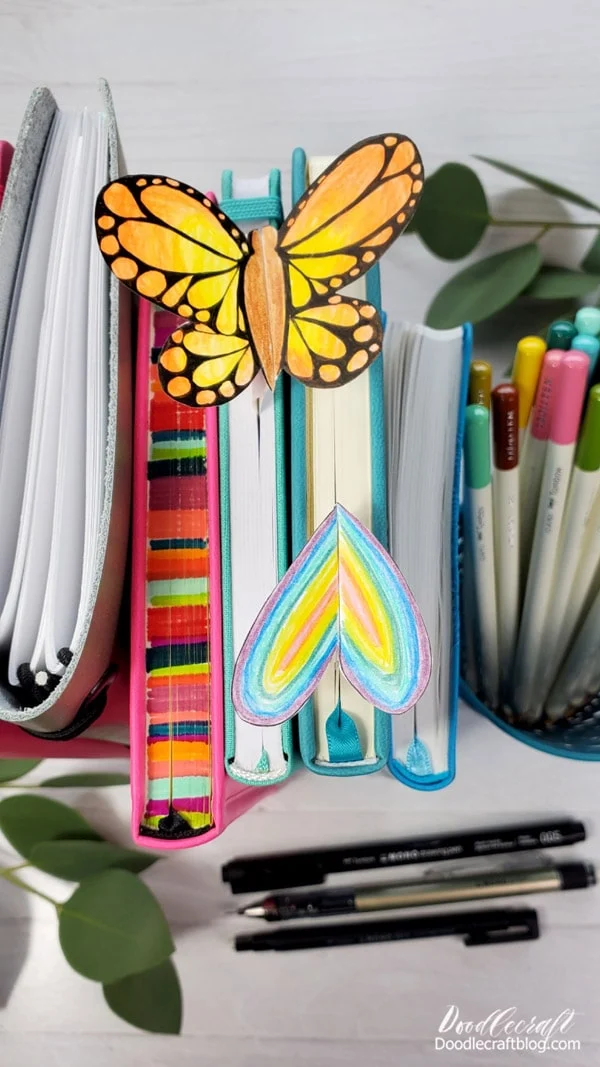 Monarch butterfly bookmark and rainbow heart bookmark peeking out of the top of notebooks on a table.