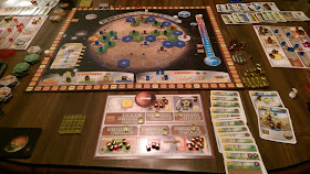 A game of Terraforming Mars in progress. The board is an image of Mars covered in a hexagonal grid with hexagonal tiles representing greenery, cities, and oceans arranged on them, and all but the ocean tiles have a coloured cube on them. There is a track from 0 to 14 represnting the atmospheric oxygen content at the top, and a thermometer running from -30º C to 8º C along the right side. A scoring track runs around the outer edge of the board. Player mats can be seen around the board, as well as tableaus of played cards and the various cubes: some in the players' colours and some in gold, silver, and bronze.