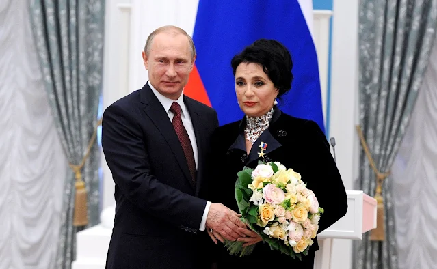 Do lado esquerdo, Valdimir Putin e do lado direito, Irina Viner. Os dois estão em uma sala branca no Kremlin e estão vestidos na cor preta. Ela está segurando um buquê de flores