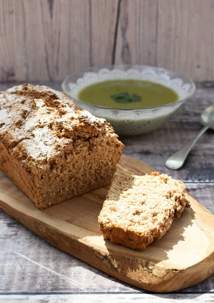 Wholemeal beer bread loaf with a slice cut on a wooden board next to a  bowl of broccoli soup