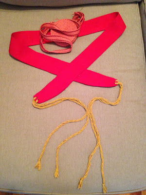 A screaming red fabric belt with gold twisted ties on the ends, laid out in a pretzel shape on a tan couch, and a roll of orange and red inkle-woven tape.