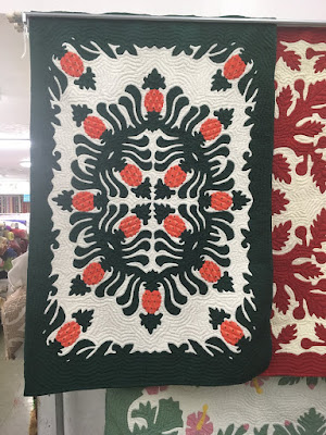 A hanging quilt with large Hawaiian applique in deep green and orange on a cream background, making a border and eight-pointed oval of pineapples, with four pineapples pointing inward at the very center.