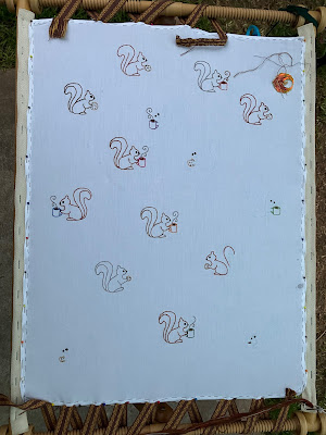 A large white rectangle of fabric has been mounted in a slate frame for embroidery. On the fabric are numerous small outlines of stylised squirrels. Half are holding coffee cups and half are holding chocolate chip cookies. Most of them have been embroidered in various colours.