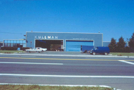 Photograph of the exterior of the Kullman building, with the company name prominently displayed above the entrance, vehicles parked in front of business.